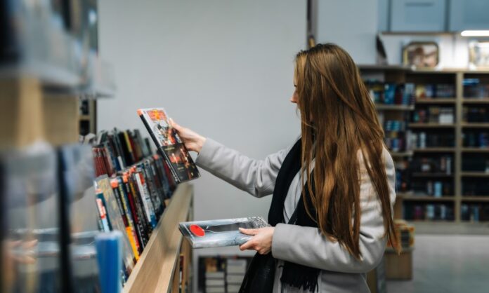 A long-haired woman browsing the graphic novel section of a book store, debating between the two novel options she holds.